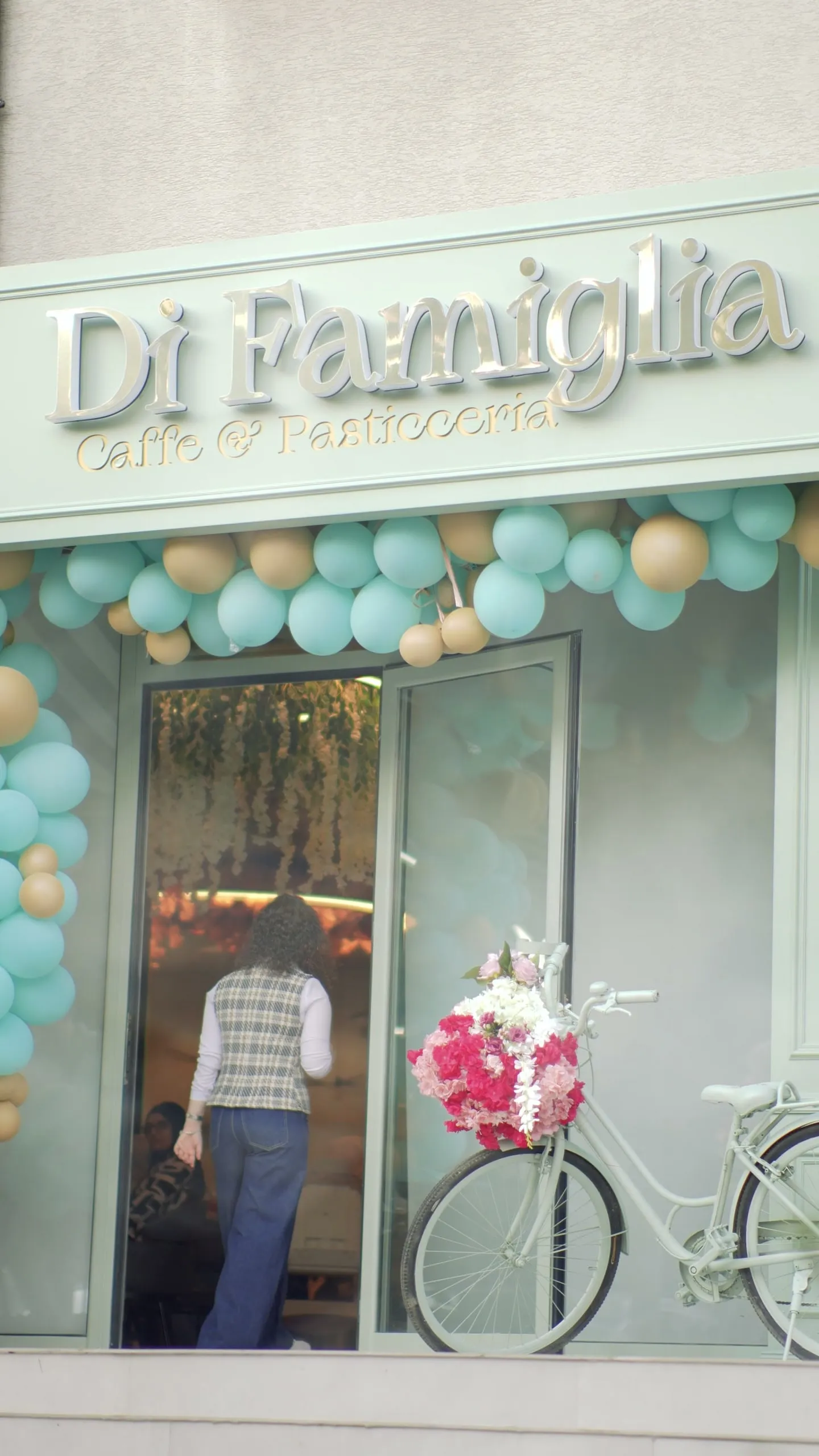 The sage-green Di Famiglia storefront on opening day: balloons over the door, a guest walking in, the white bicycle with flowers